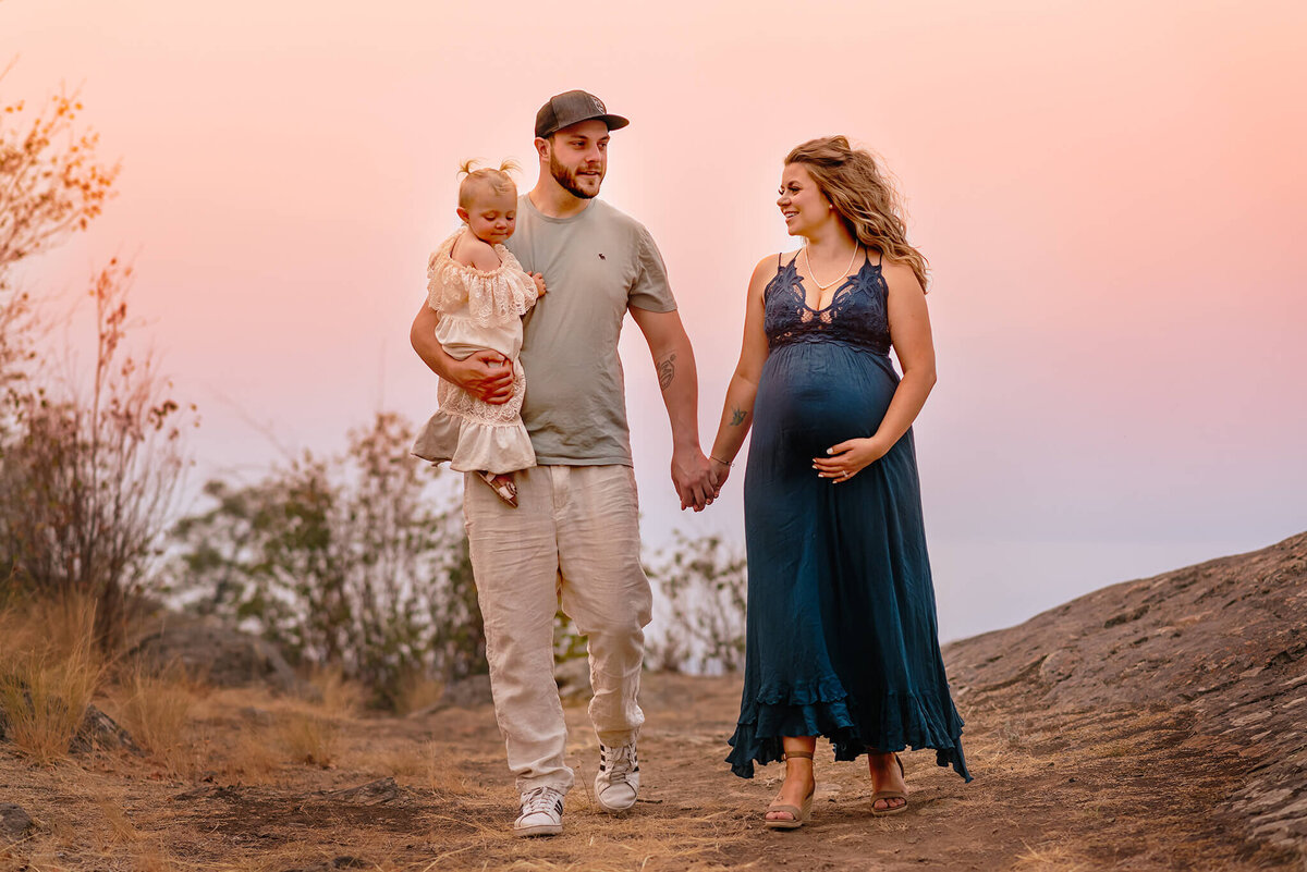 A small family holds hands while walking with a sunset background. Husband and wife smile at eachother while the woman holds her pregnant belly.Taken in Tobiano, Kamloops by Brittany Danielle