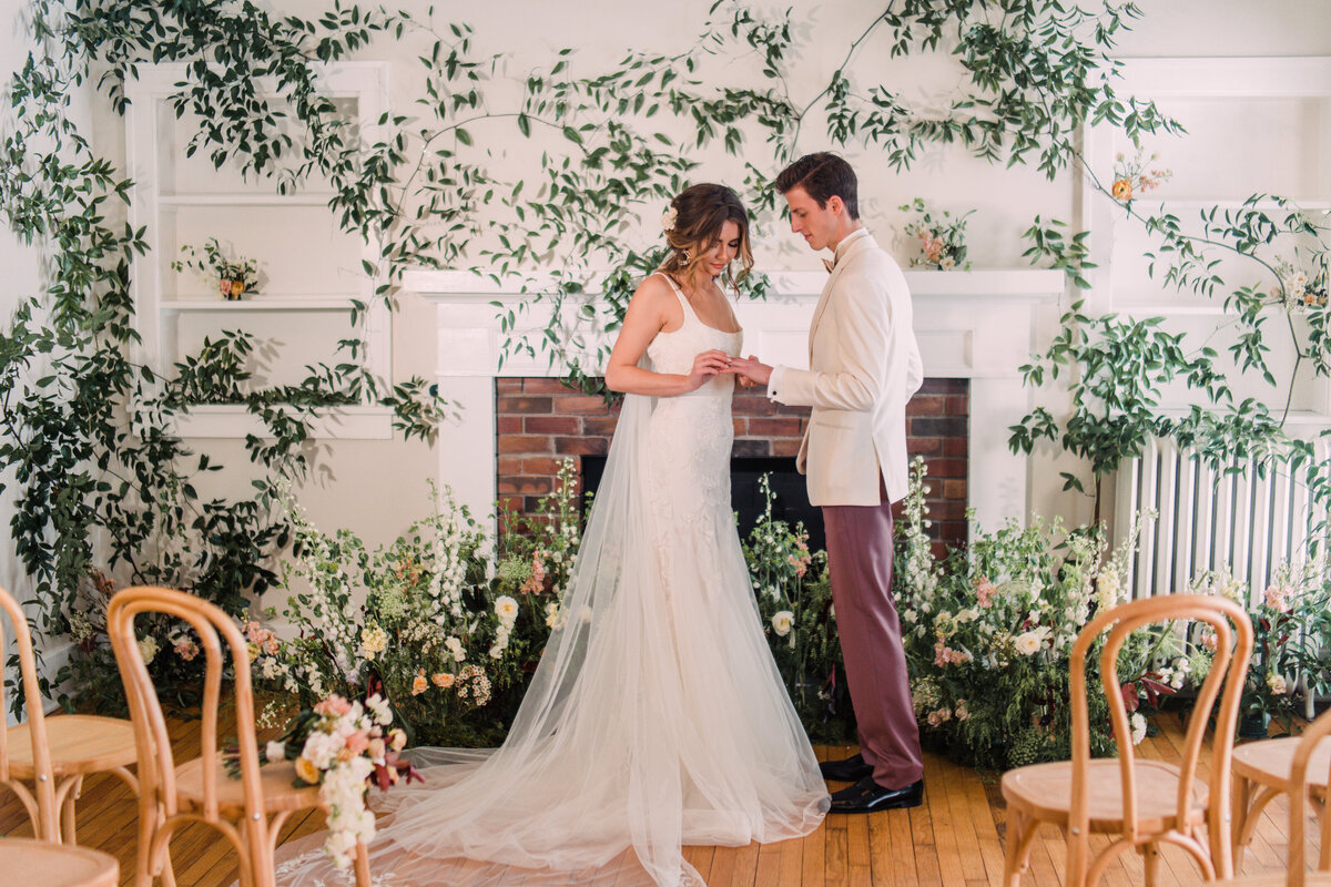 Bride wearing Alexandra Grecco exchanging rings with groom in white & rose tuxedo in a room decorated with vines & flowers