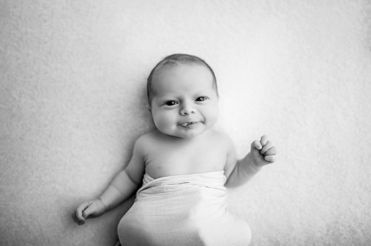 Black and white portrait of newborn baby staring at camera blowing spit bubbles while swaddled in white muslin wrap.