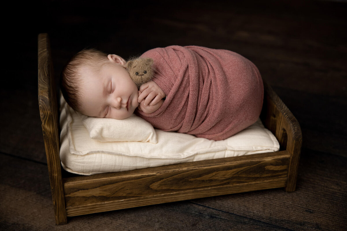 newborn baby asleep on her side wrapped in a pink fabric on a tiny wooden bed holding a tiny teddy bear