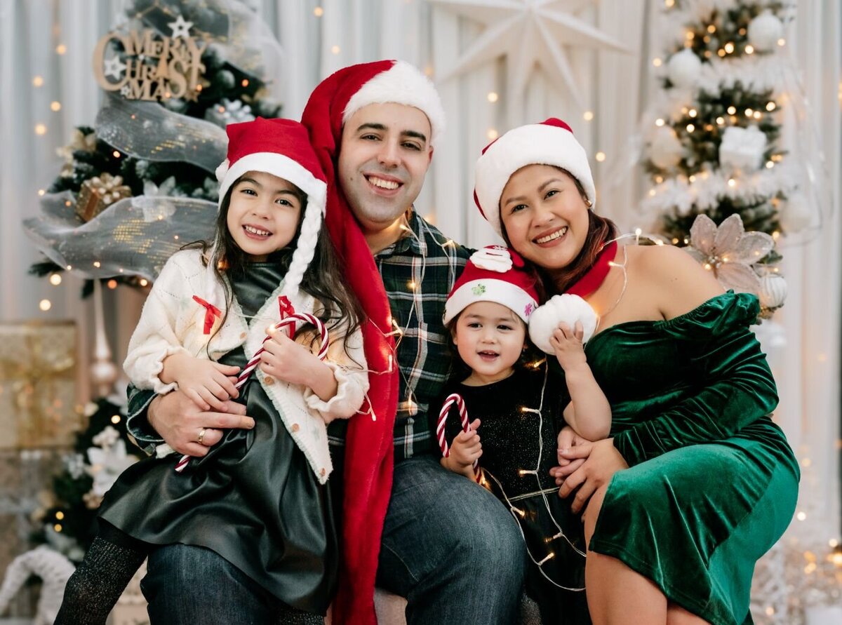 Smiling family of four wearing Santa hats and holding candy canes in a cozy Christmas photo studio.