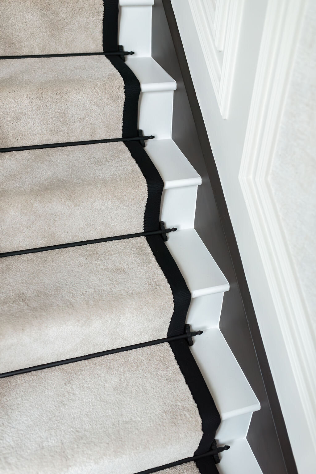 Close view of a a white staircase featuring a beige runner bordered by black trim against bright white walls.