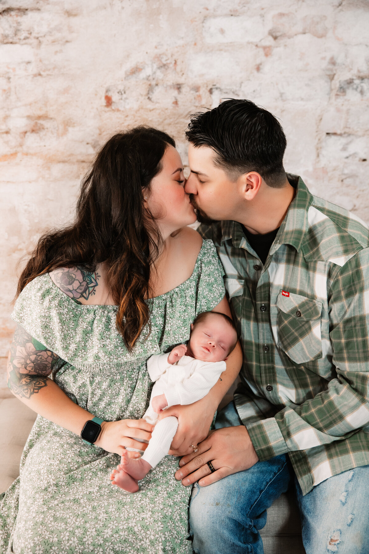 Happy new parents in green kiss while their newborn baby sleeps in mom's arms on a bench in a studio of a Milwaukee Newborn Photographer
