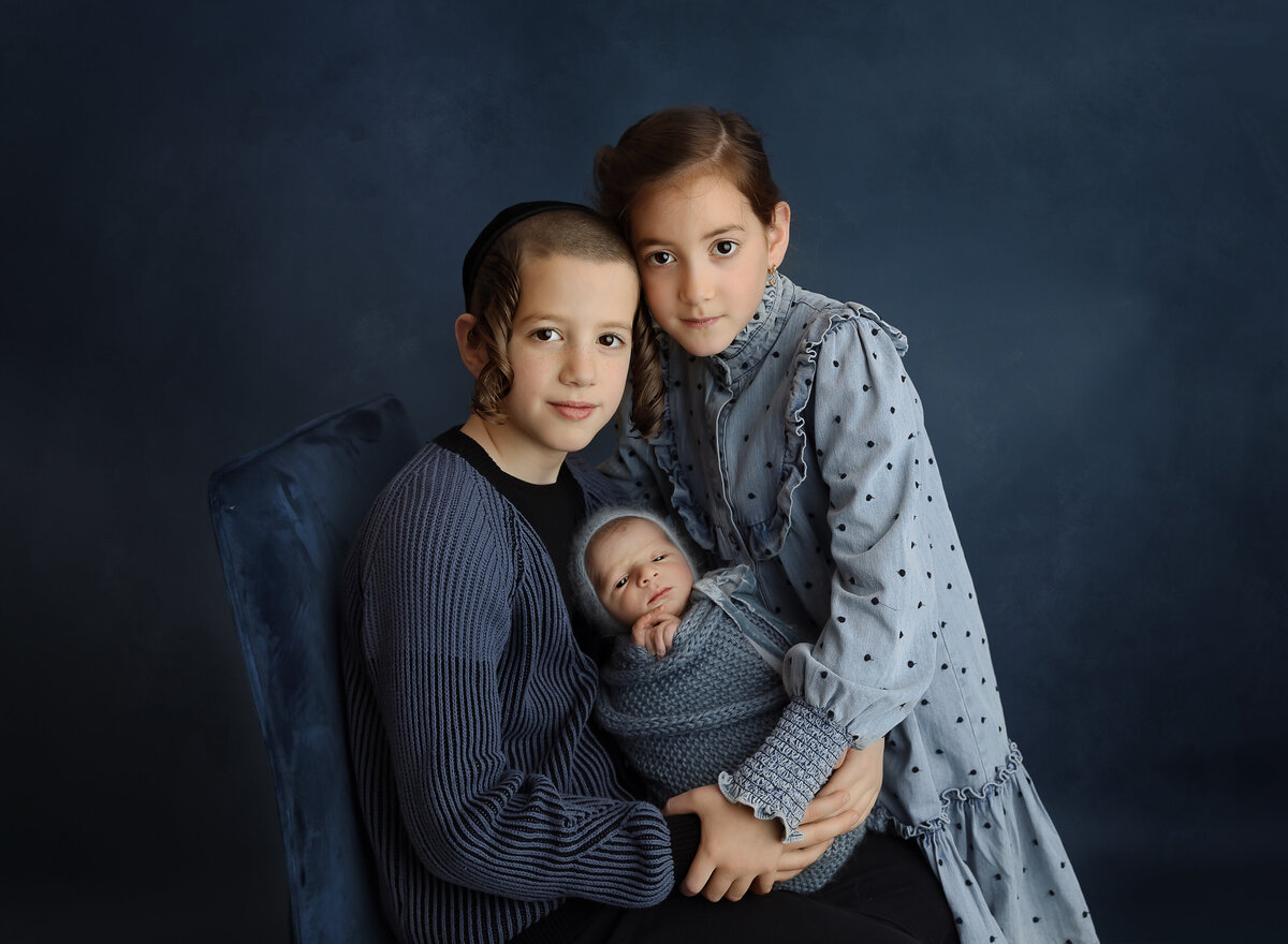 Brother and sister hold their newborn baby brother between them for a newborn photoshoot in brooklyn, ny. Brother and sister's heads are resting together and smiling at the camera. Captured by top Brooklyn newborn photographer Chaya Bornstein