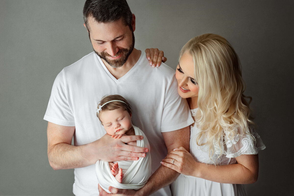 new parents wearing white holding their newborn baby on a grey   backdrop