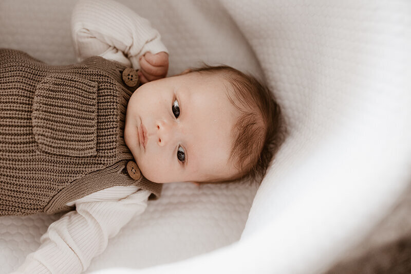 Newborn baby boy in a knit overalls set while laying in a white lined Moses basket.