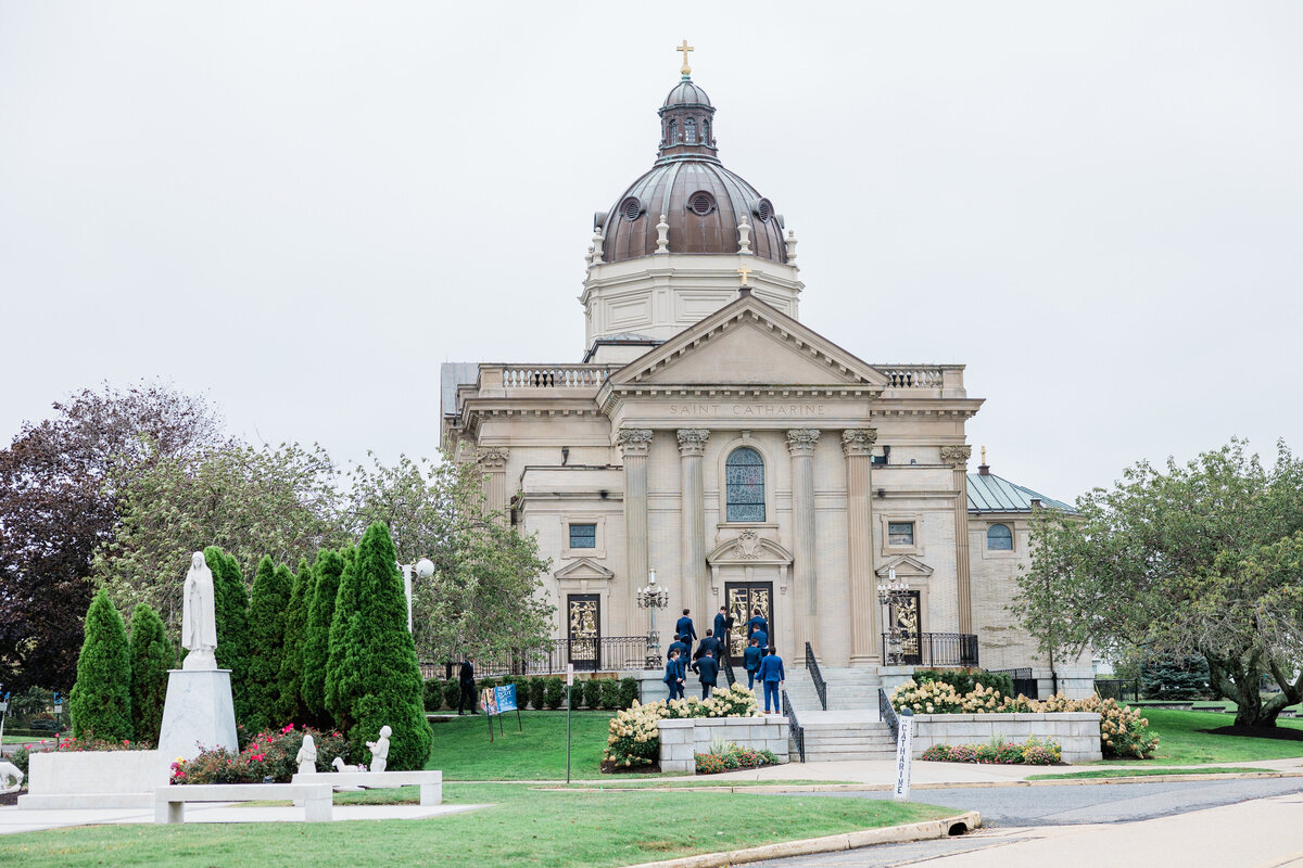  A grand, historic church-style venue with a domed roof and classical architecture. Guests in suits and dresses walk up the steps, ready to celebrate the couple’s wedding day.
