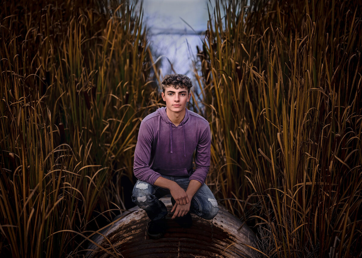 A high school senior squats in a field of tall grasses in a purple hoodie and jeans for his Pella Senior Photographer