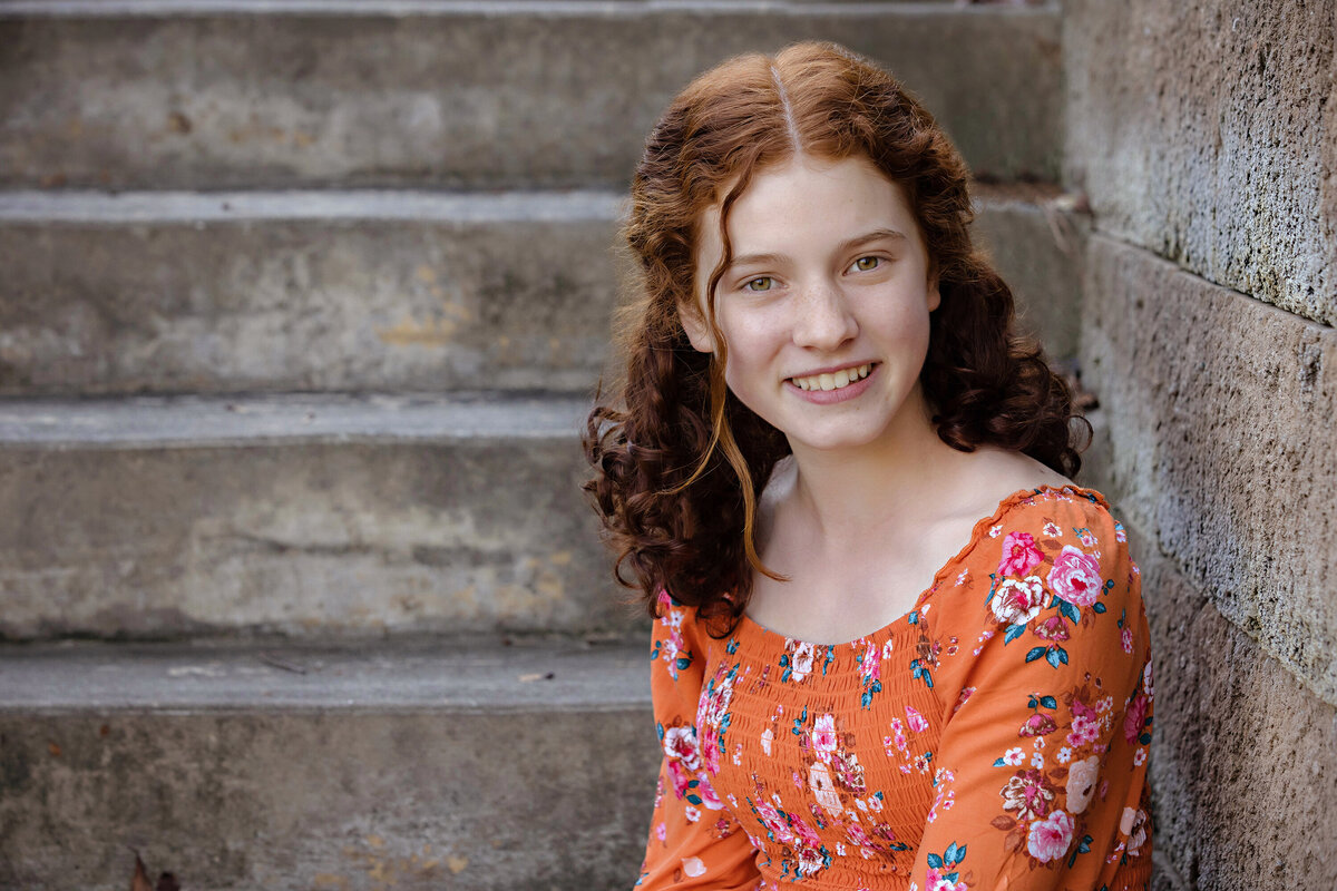 Radiant red‑haired teen in orange floral dress seated on textured concrete staircase at Ballantyne Corporate Park Charlotte NC