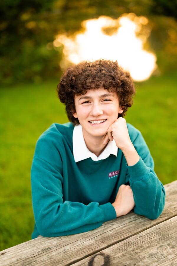 High school senior boy smiling at picnic table with green background.