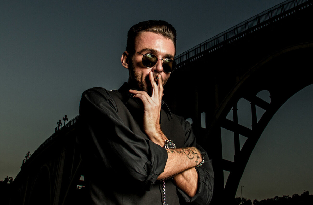 Portrait of musician Chandler Morrison smoking with arms folded while standing beneath bridge