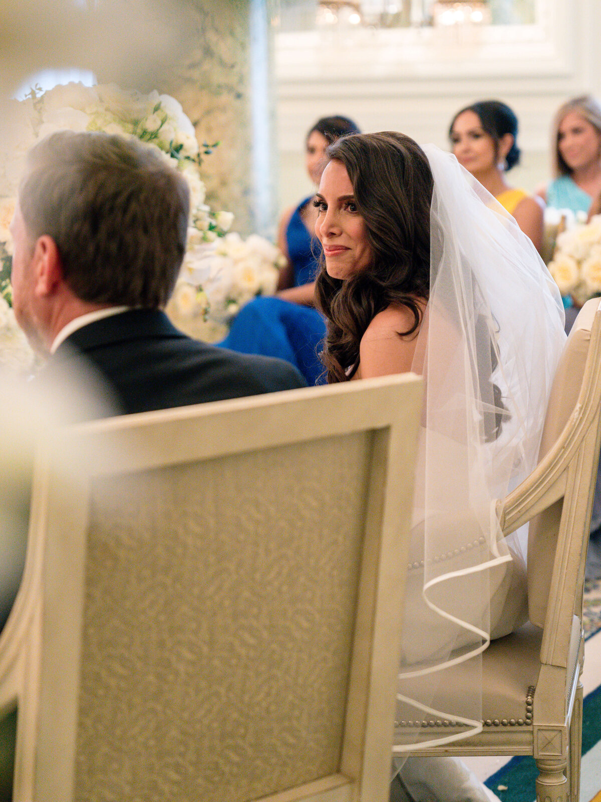 Bride in a white dress and veil smiles while seated during a wedding ceremony. She is surrounded by bridesmaids in blue dresses. The setting is elegantly decorated with white flowers and soft lighting.