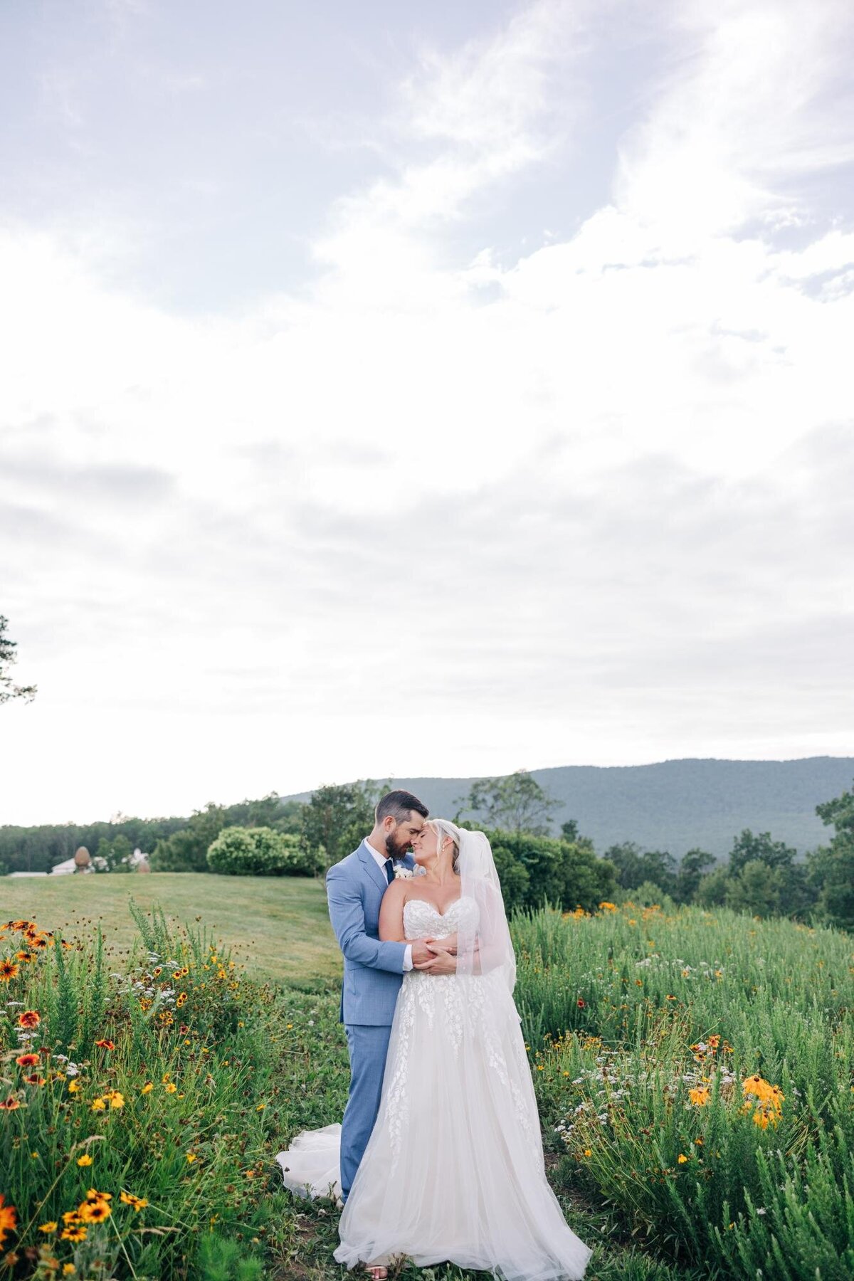 A couple in wedding attire embracing in a field with wildflowers, under a cloudy sky.