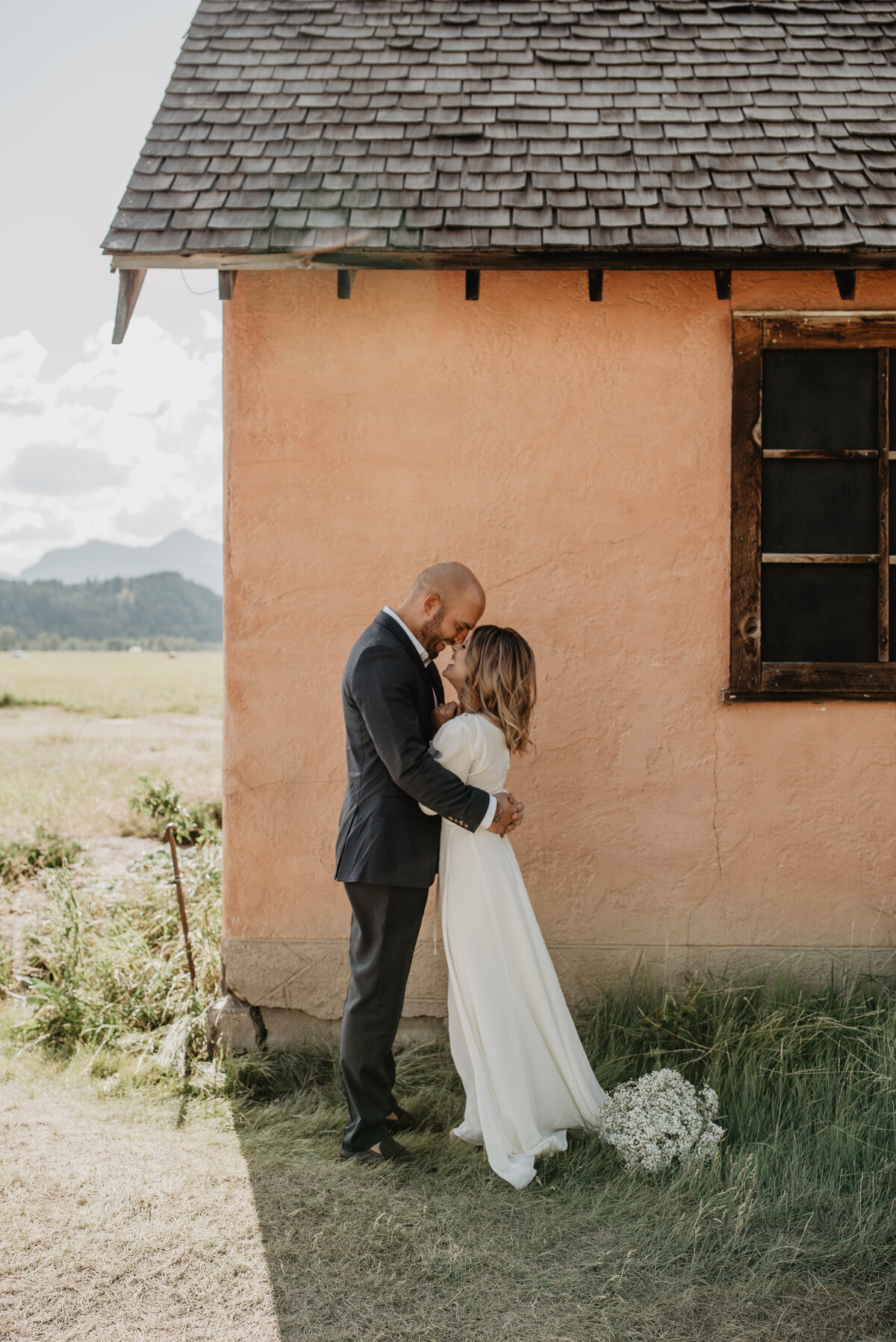 jackson hole wedding day bridals with bride and groom standing in front of an old rustic house with the groom kissing the bride's forehead and holding her waist