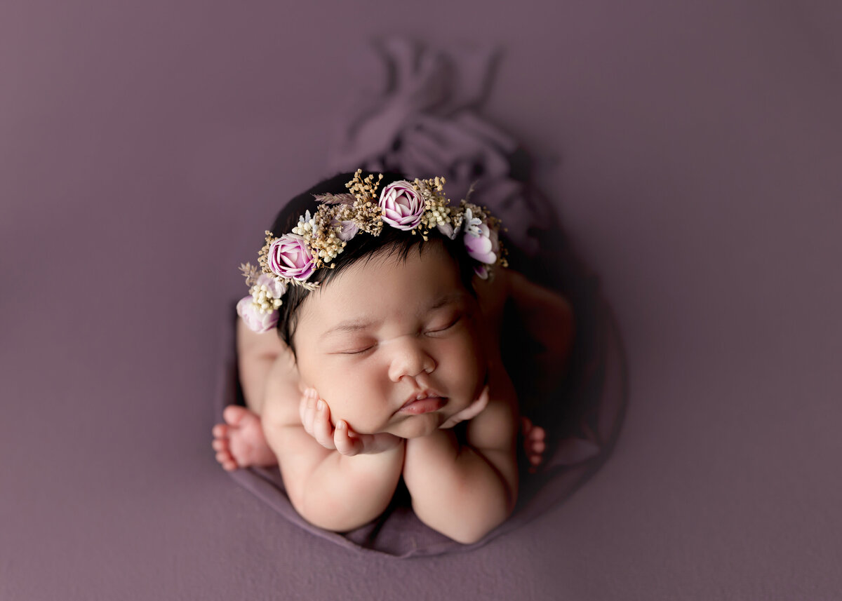 Baby girl photoshoot in West Palm Beach and Boynton Beach Florida studio.  Baby is in froggy pose, resting her hands under her chin and propped up on her elbows. Baby is atop of a purple stretch fabric wearing a purple and moss green floral crown.