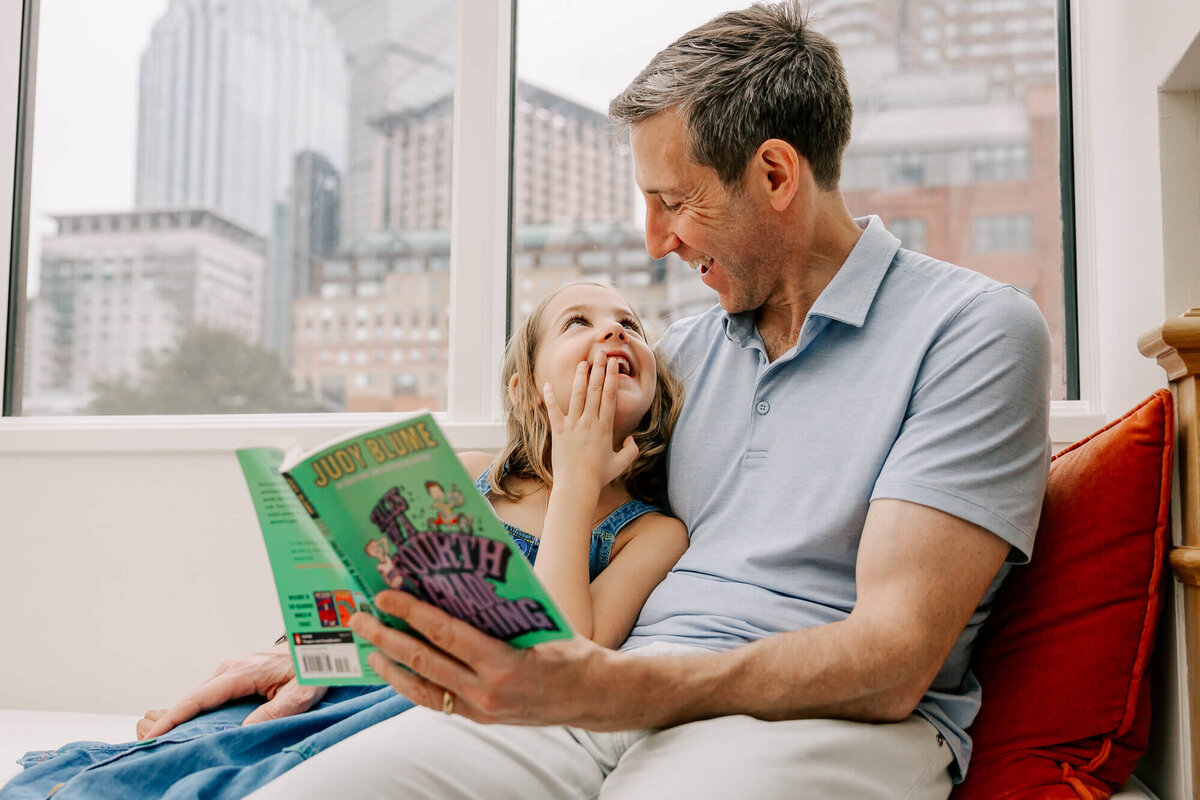 Dad and young daughter snuggle in their South End, Boston bay window, reading "Tales of a Fourth Grade Nothing" together. The Boston skyline is behind them.