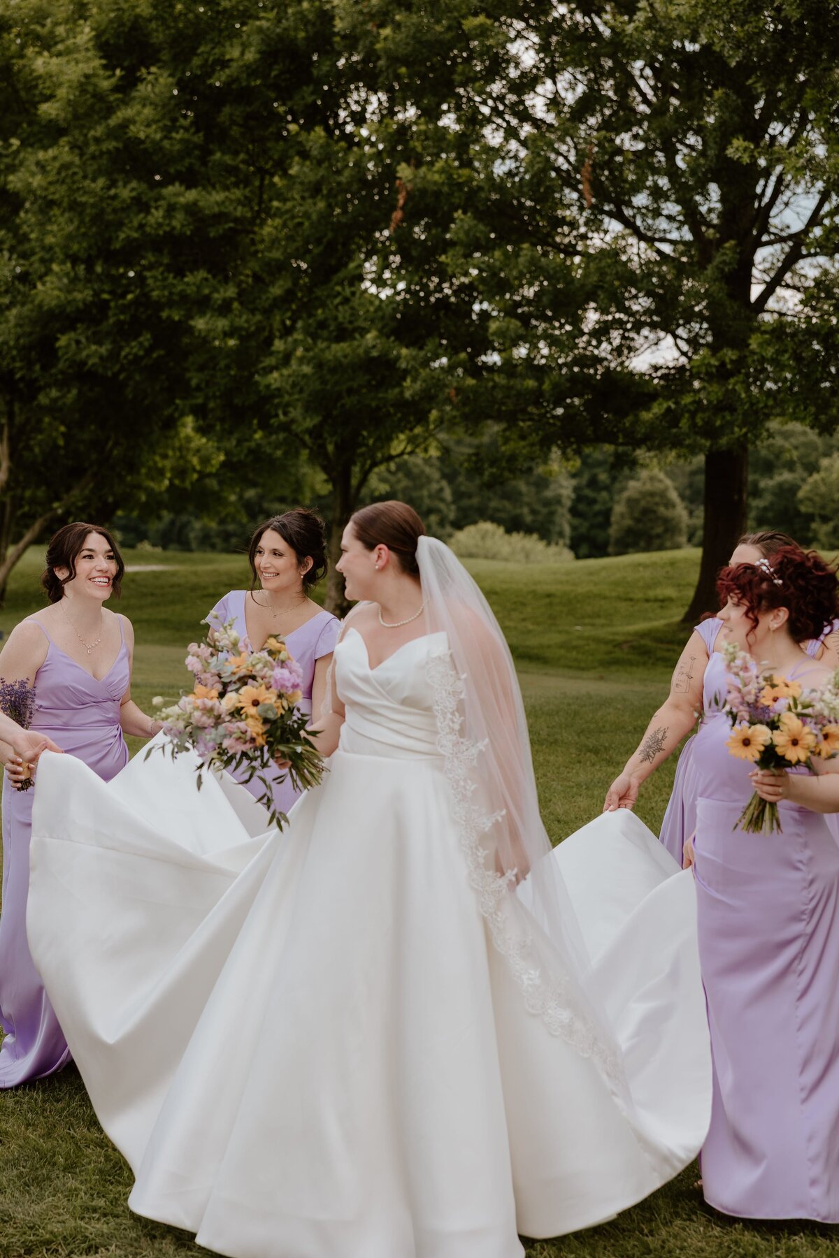 Bride surrounded by bridesmaids in lavender dresses, all smiling and holding bouquets, as they help arrange her gown outdoors on a lush green lawn.