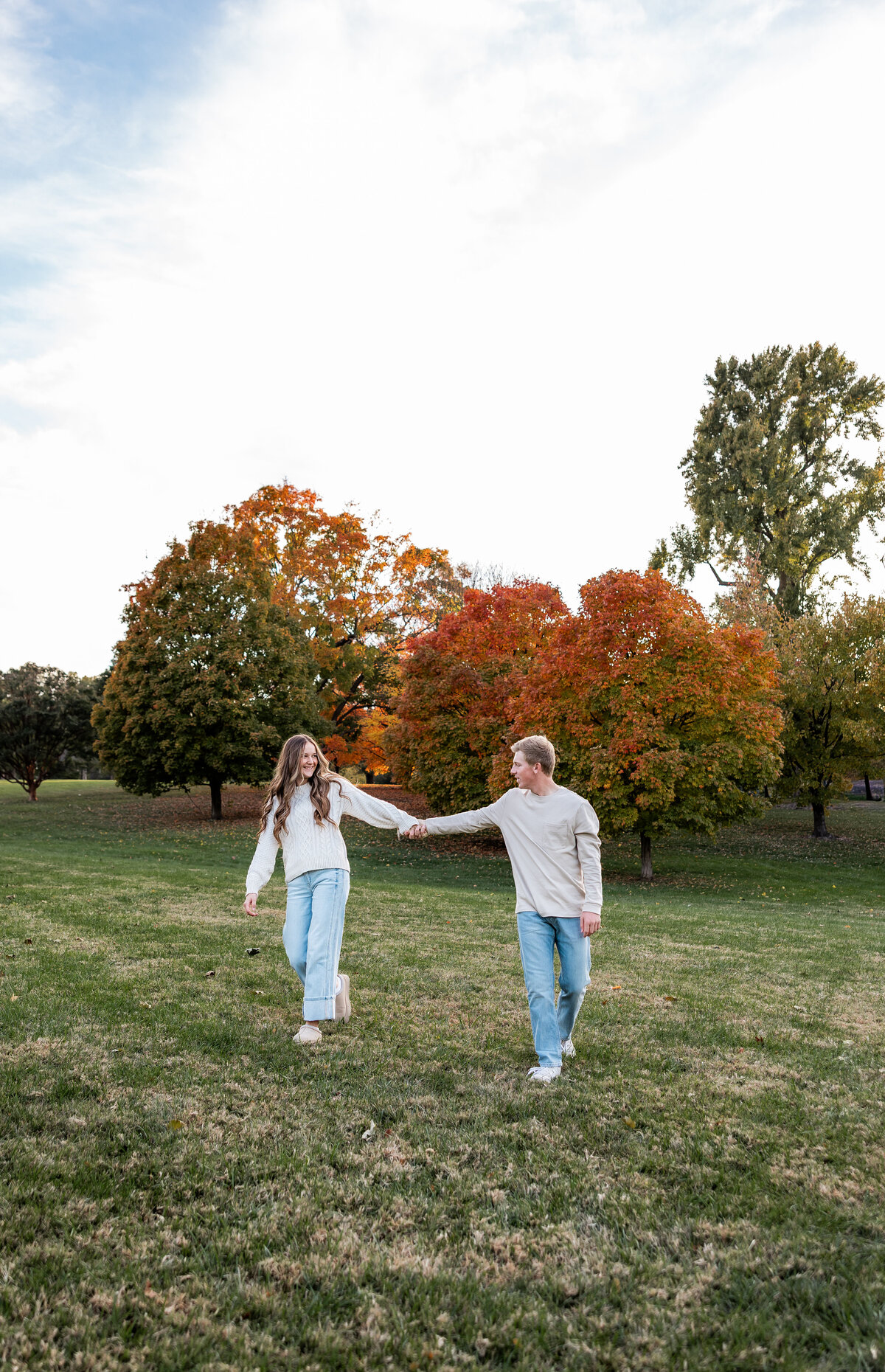 A young couple wearing cozy fall outfits walk hand in hand in a field at Loose Park in Kansas City with fall leaves in the background.