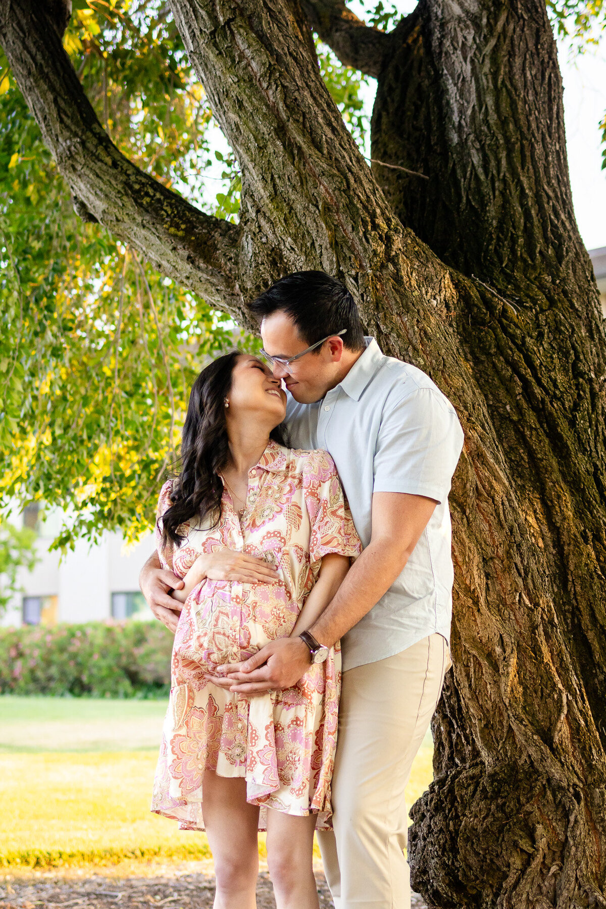 Couple shares a tender moment leaning into one another beneath a lush tree during their maternity session — Bay Area Maternity Portfolio - Ellobelle Photography.