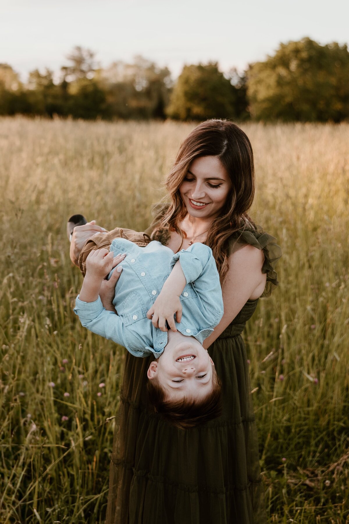 A smiling mother holding her young child upside down in a sunlit field, captured by a Hudson Valley family photographer.