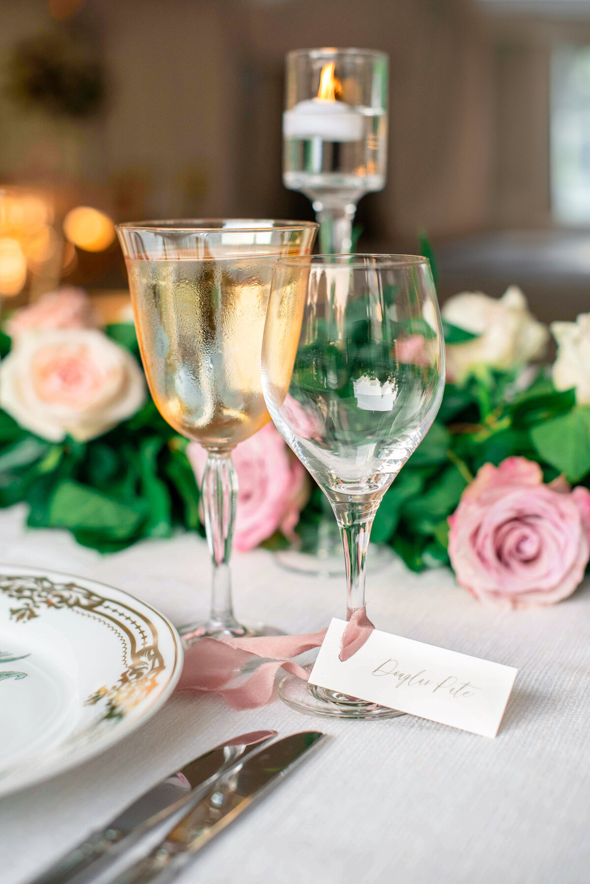 A table setting featuring a filled wine glass, an empty wine glass, and a decorative plate with a floral design. Pink roses and greenery are in the background, with a lit candle in a tall holder.