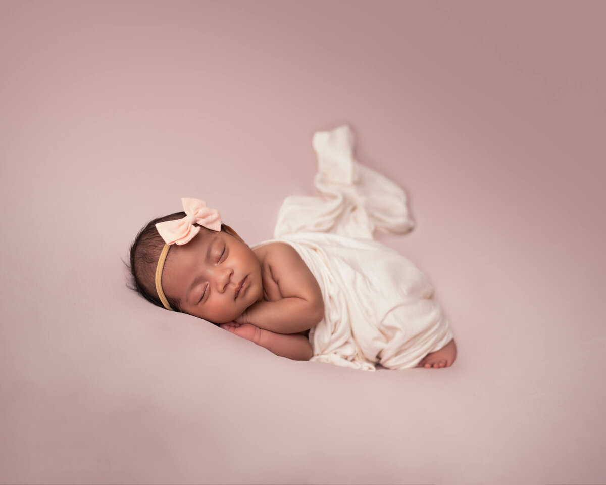 a newborn baby girl wrapped in pale pink and wearing a large pink bow sleeps with her hands under her cheeks