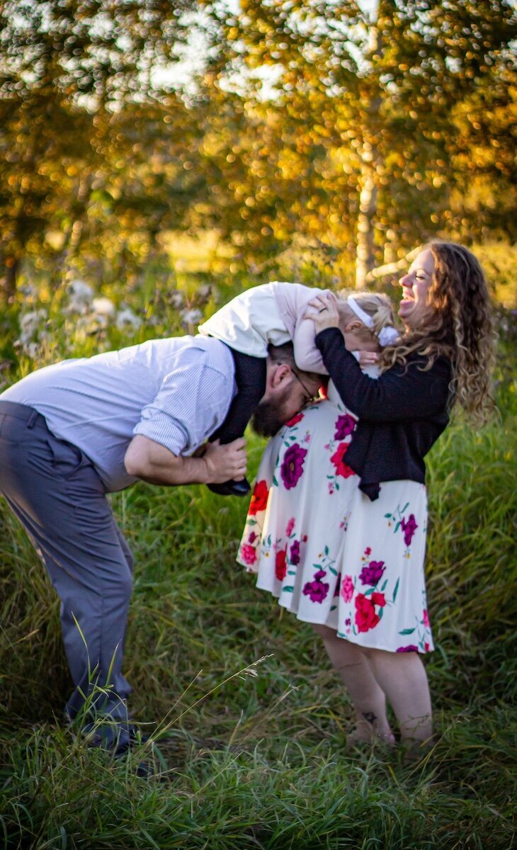 a dad has his toddler daughter on his shoulders leaning forward and kissing his pregnant wife's belly