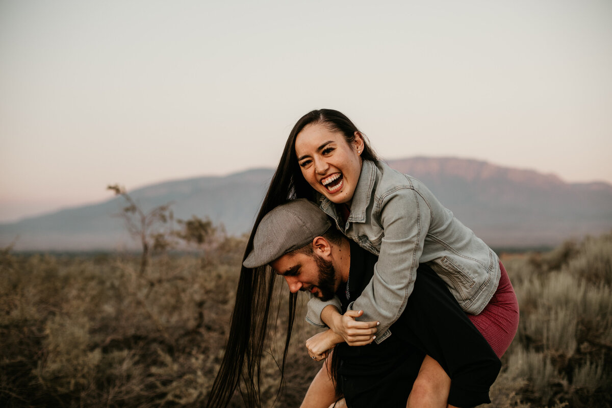 woman laughing while on fiancé's back in desert