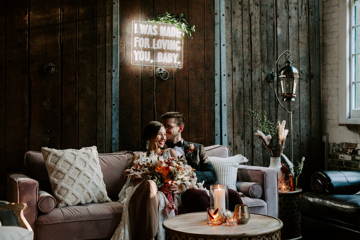 Bride and groom on a couch under a neon sign that reads "I was made for loving you"