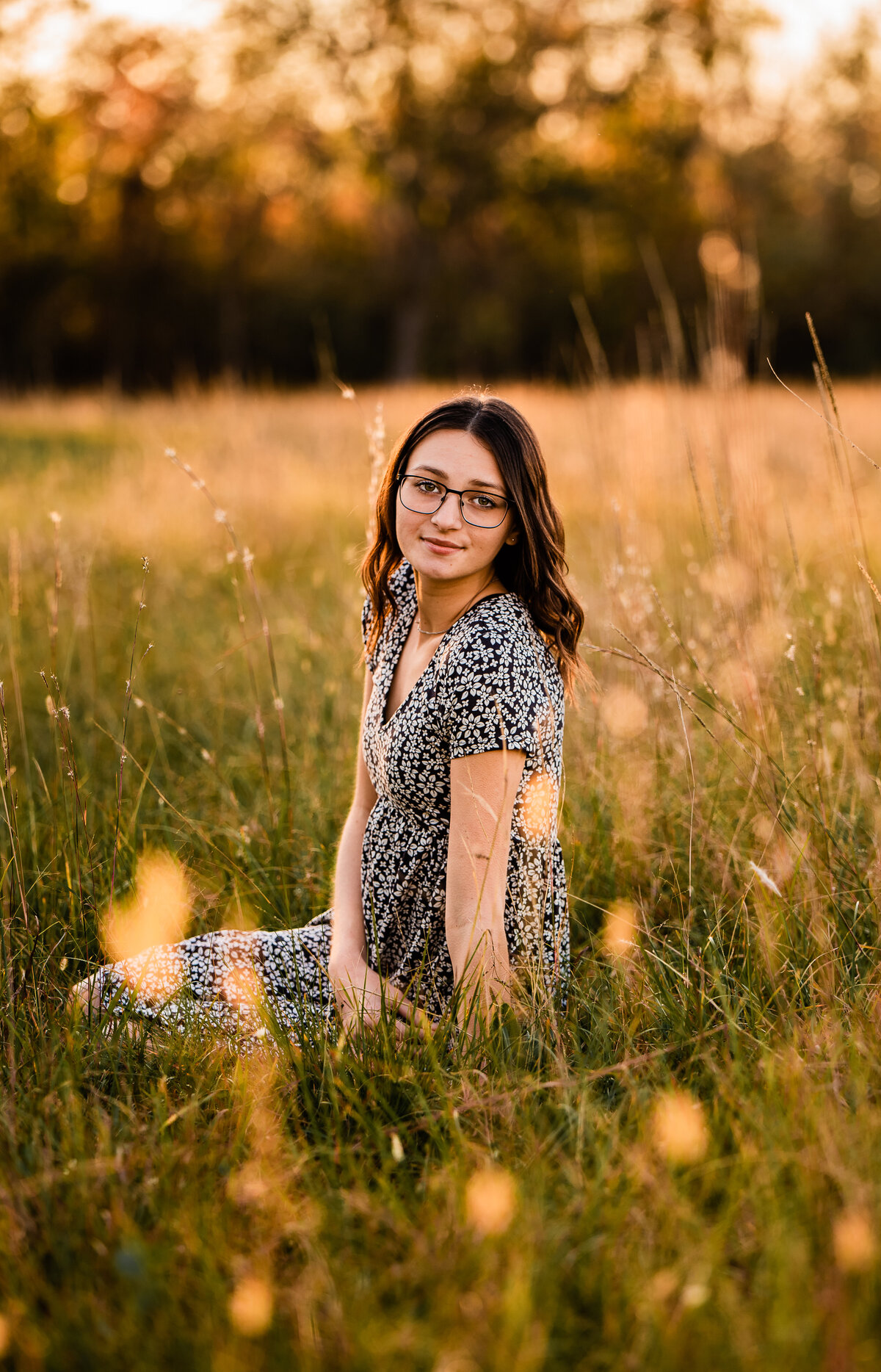A girl graduate sits in a field of golden grass at sunset and smiles at the camera.