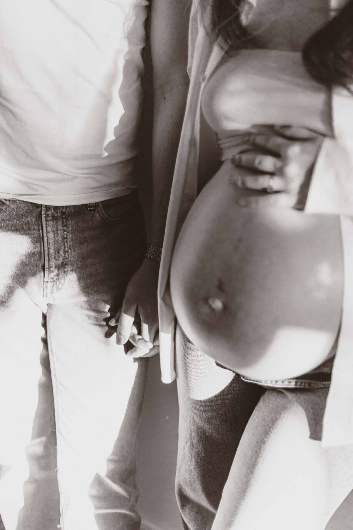 En svartvit närbild på en gravid kvinnas mage och hennes partner hållandes hand med vardagliga kläder på sig