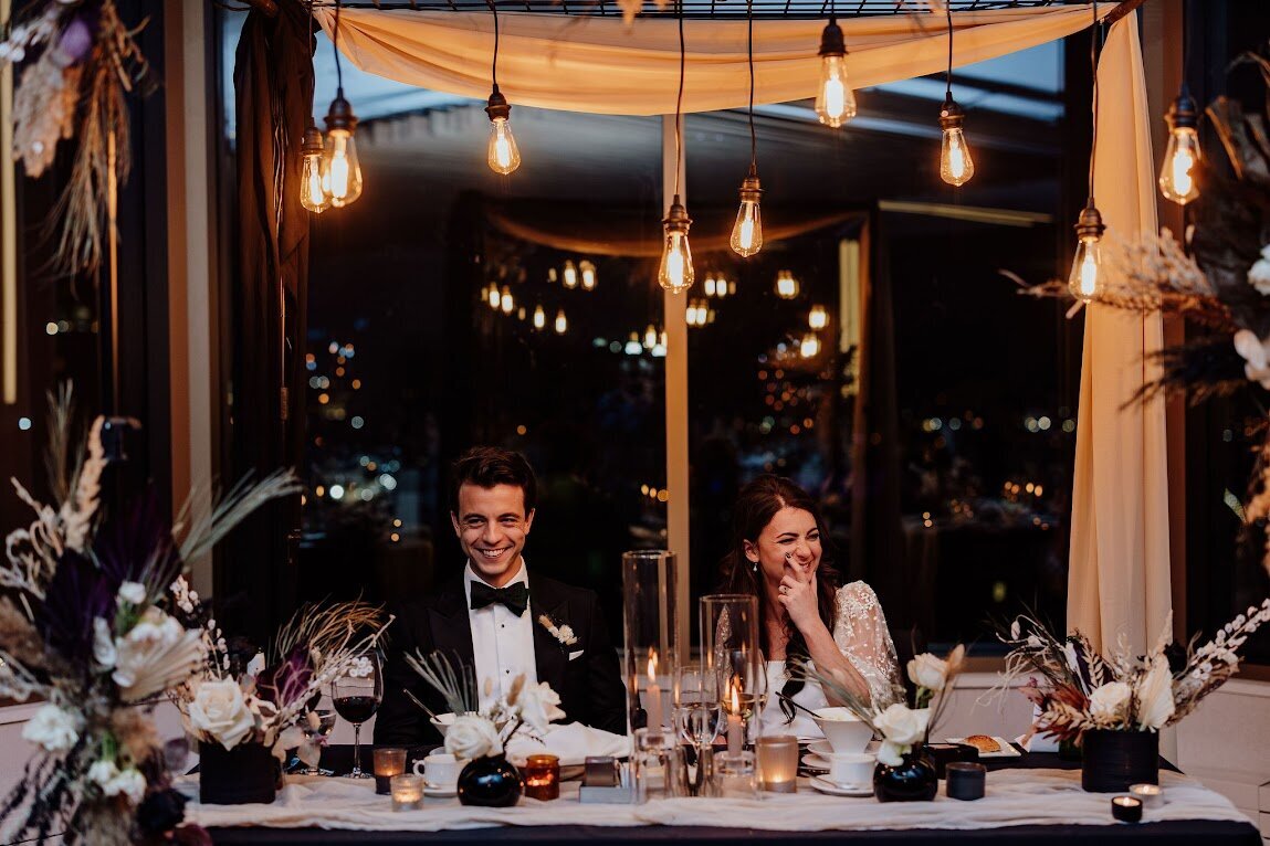 A rock-star couple sit at their sweetheart table made of industrial lights, dried white and black flowers and creative draping over a metal frame  at their wedding at the venue TwentyTwo in the Westin Hotel in Ottawa Ontario