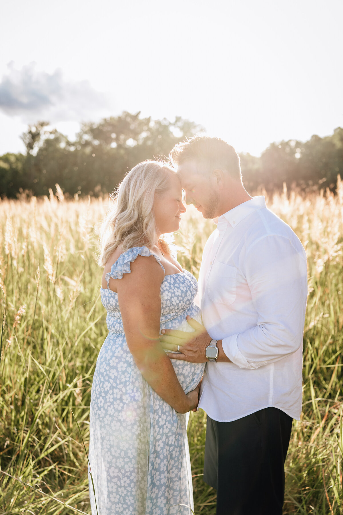 A pregnant couple stands closely in a sunlit field, touching foreheads and cradling the woman’s belly together, both smiling gently.