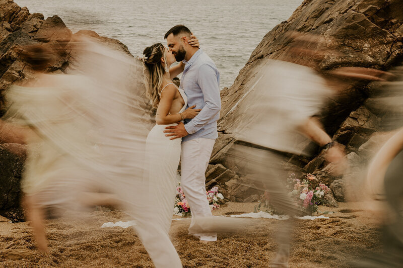 Couple s'embrassant dans un décor de rochers et de plage entourés d'amis en mouvement donnant un effet de flou. Séance photo en Vendée.