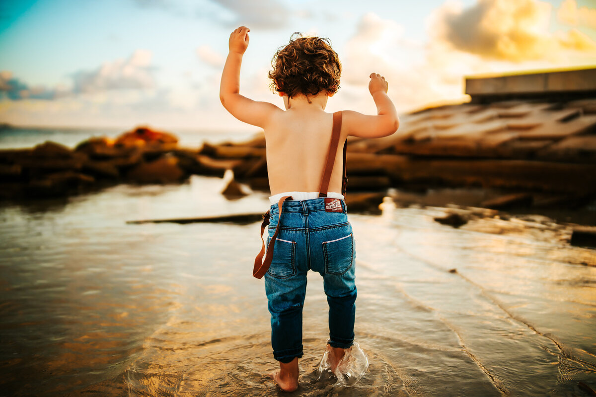 colorful sunset beach portrait of a toddler in jeans and suspender playing in the water