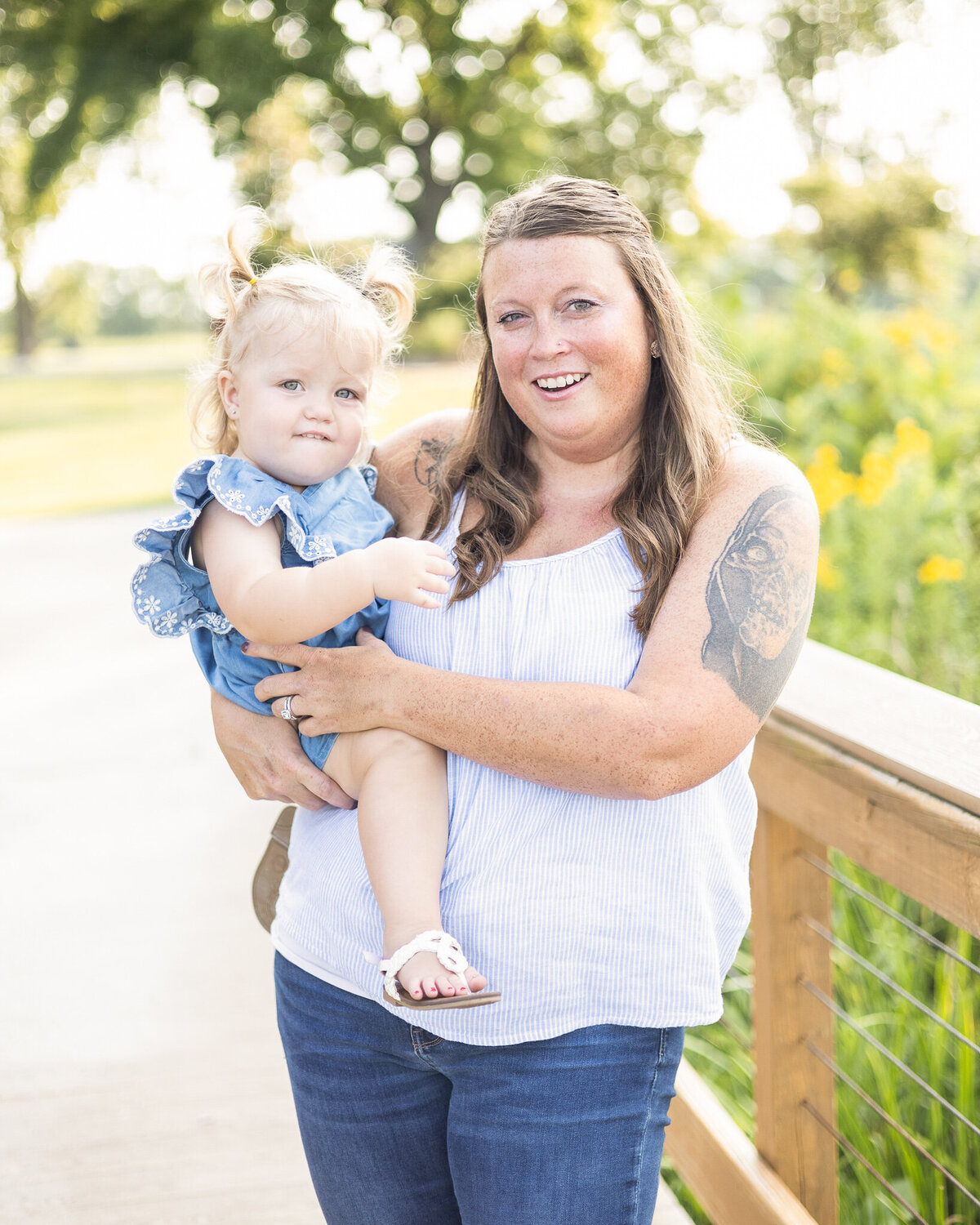 Mom and toddler daughter standing on a bridge at the wildlife sanctuary,has long brown hair, wearing a sleeveless white shirt, blue jeans, and brown sandals, smiling at the camera.  Toddler daughter is in her arms with blonde pigtails, blue dress, looking at the camera.