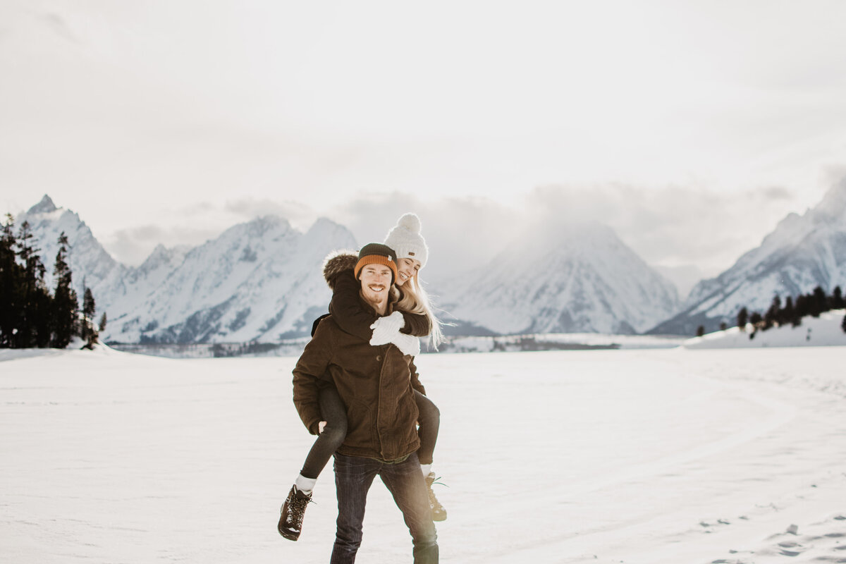 man holding woman on his back in a snowy field in front of the Grand Tetons while smiling during their winter engagement session captured by jackson hole photographers