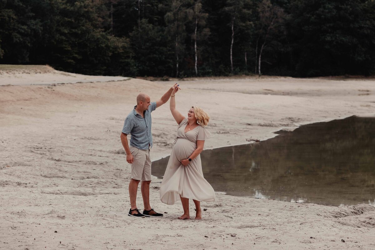 Een man en een vrouw op het strand, zij is zwanger en houd haar buik vast en ze dansen samen tijdens de zwangerschapsshoot in Zwartsluis