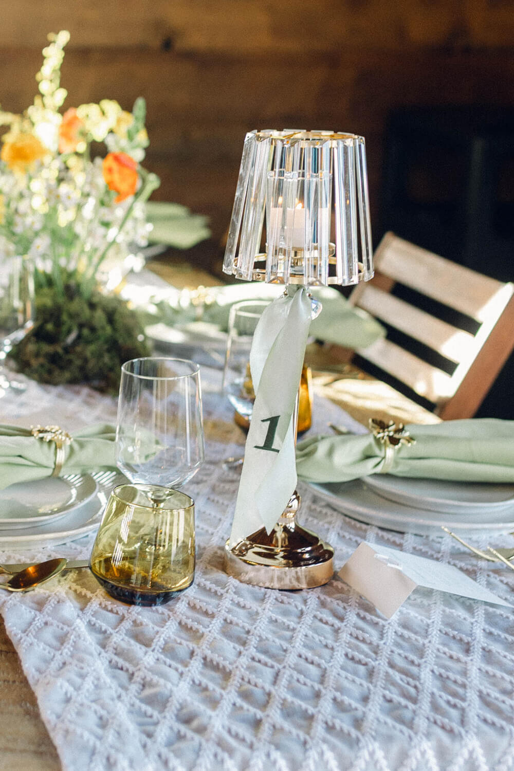 Embroidered ribbon table number tied around a crystal lamp centerpiece, set on a lace runner with sage green napkins and amber glassware.