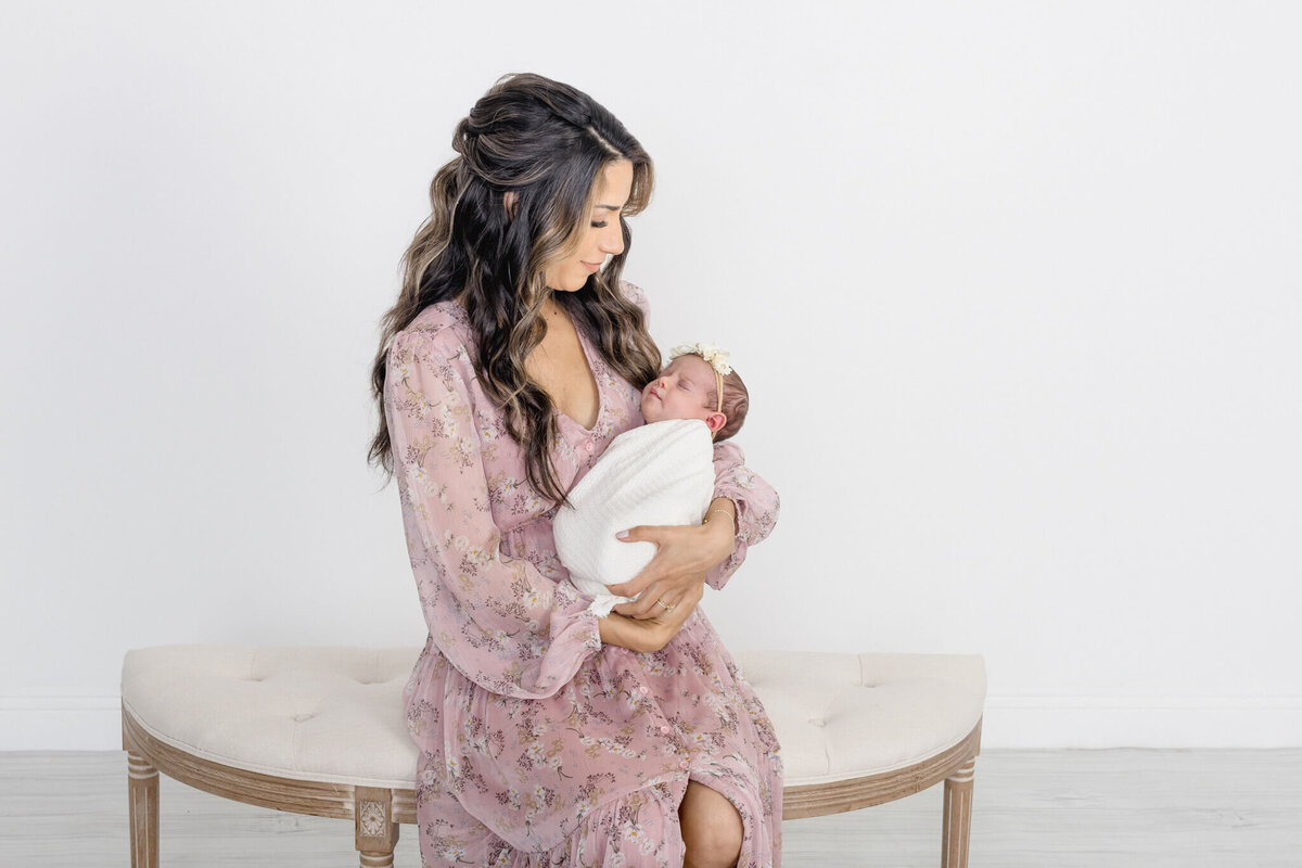 A woman in a light pink floral dress sits on a cushioned bench, gently cradling a newborn wrapped in a white blanket. The scene is set against a plain white background.