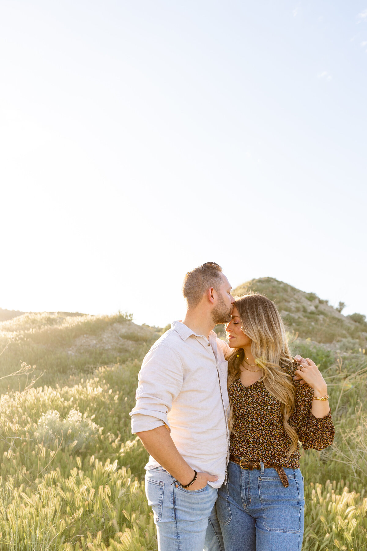 A man gently kisses his fiancée’s forehead as they stand together in a grassy field in Saratoga Springs, Utah. The golden sunlight highlights their intimate moment.
