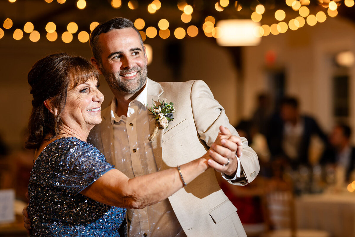 The groom shares a warm, affectionate dance with an older woman in a sparkling blue dress at the wedding reception.