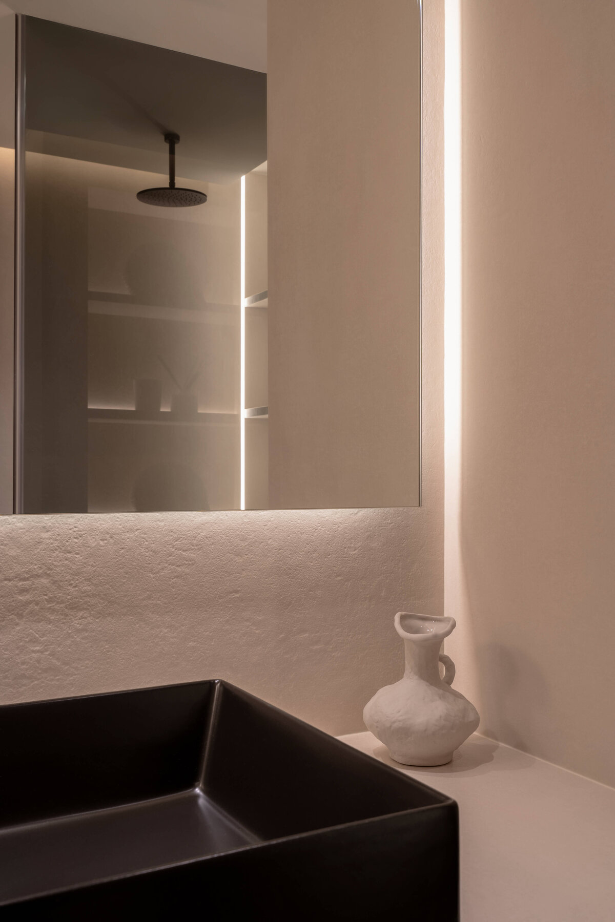 A modern bathroom with a focus on light, neutral tones. A stark black sink adds contrast and intrigue to the space. Smokey glass of a shower can be seen in the mirror.