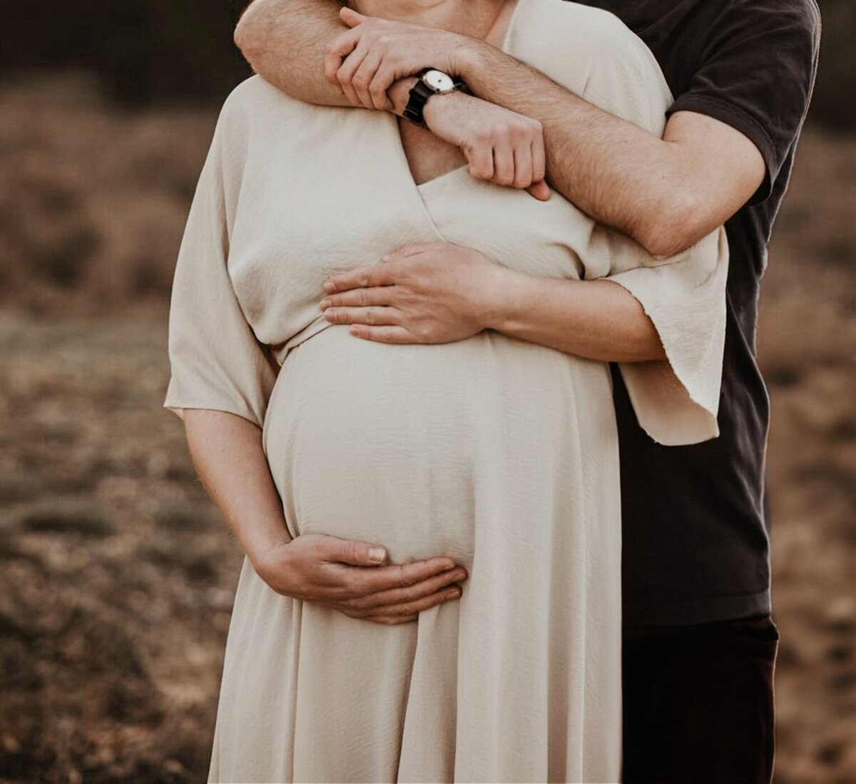 Een close-up van een zwangere vrouw die omhelsd wordt door haar partner, met de focus op hun handen op haar buik