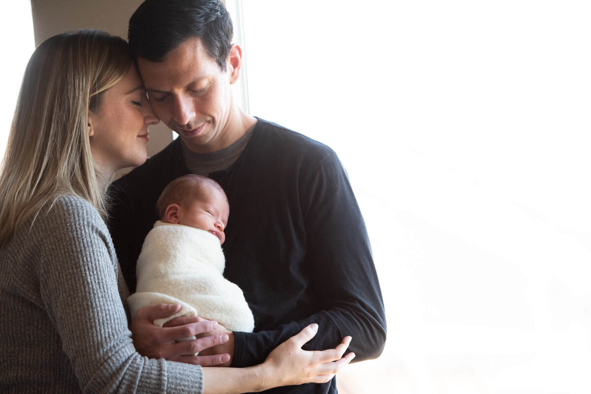 Happy new parents hug while cradling their sleeping newborn baby in a white swaddle