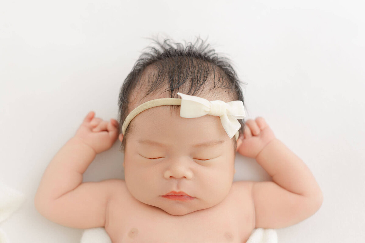 Baby girl sleeping on back with hands up next to head and laying on a minimalist white backdrop.