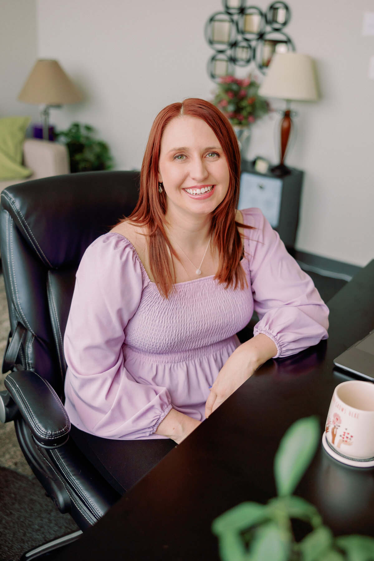 A person smiles in a purple shirt while leaning against a desk.