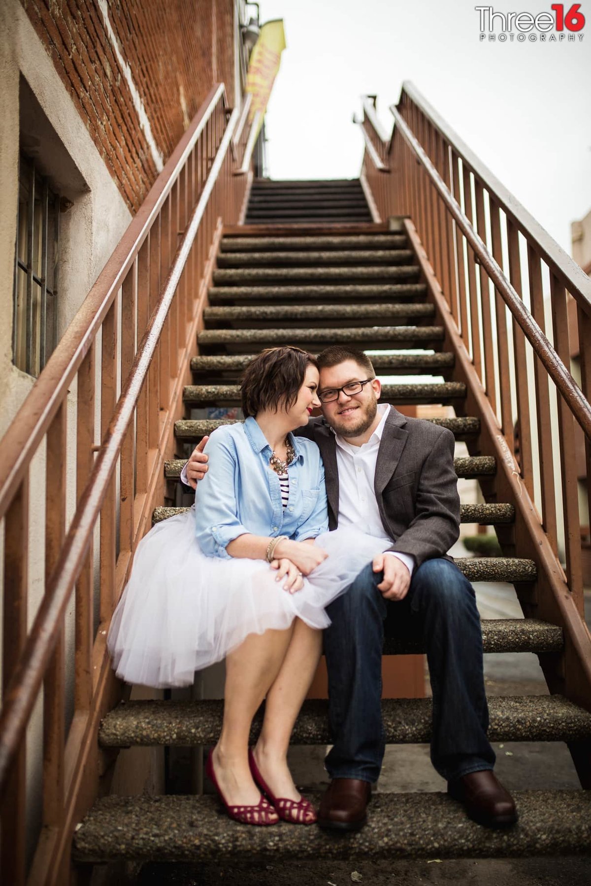 Engaged couple cuddle up as they sit together on an outdoor staircase