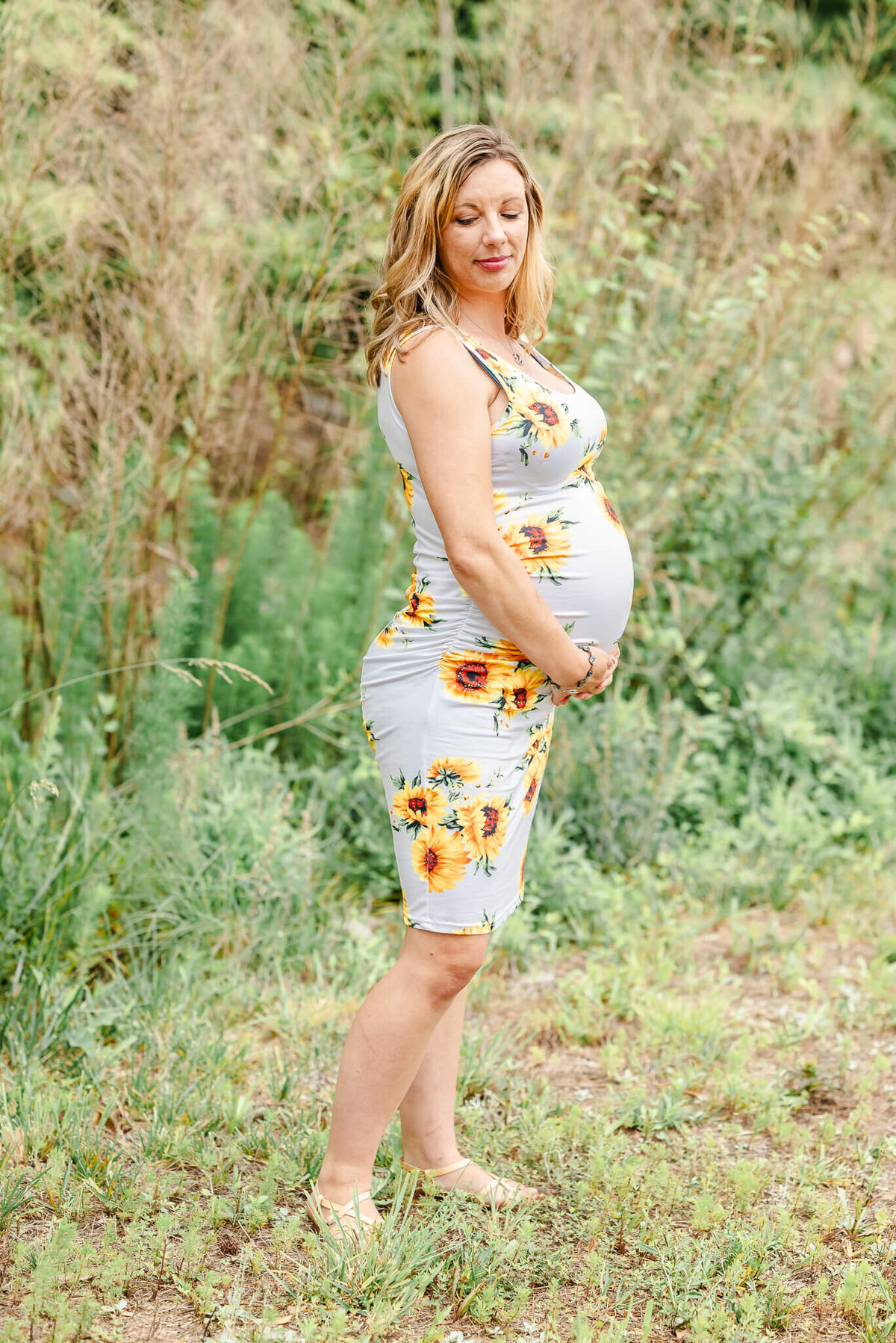 A pregnant woman, wearing a bodycon dress with sunflowers, lovingly holds her growing belly in a session with Justine Renee Photography.