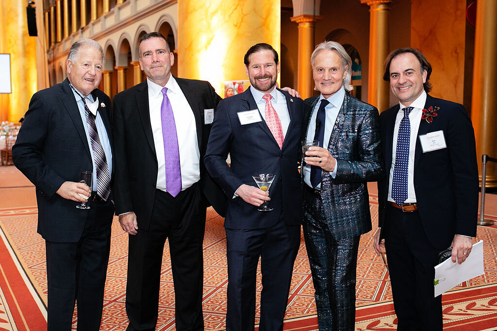 Five people pose in suits indoors.